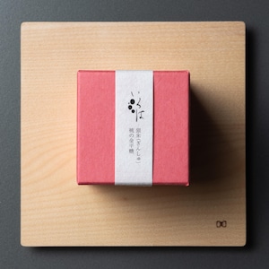 May include: A pink gift box with Japanese text on a white band, placed on a light wooden board. The box is centrally positioned on the board, which is set against a dark grey background. The box is square in shape.