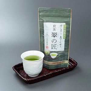 Könnte beinhalten: Eine weiße Keramiktasse mit grünem Tee steht auf einem dunkelroten, rechteckigen geflochtenen Tablett neben einem vertikalen grünen Beutel mit Premium-Grüntee. Der Beutel zeigt japanischen Text, eine kleine Grafik von Teetassen und die Aufschrift Premium Green Tea Tea Bag 16P.