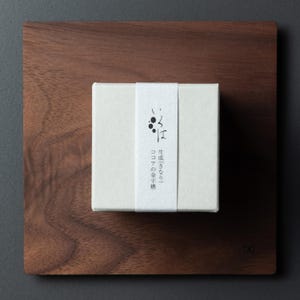 May include: An off-white box with Japanese text and a black graphic on a white band, set on a dark wooden square. The box is centrally positioned on the wooden surface, offering a minimalist design.