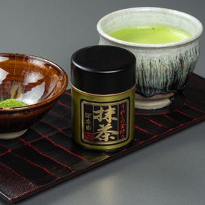 May include: A gold and black matcha tea tin with Japanese text on a black tray with a red grid pattern. A bowl of matcha powder and a ceramic bowl of green matcha tea are also on the tray.