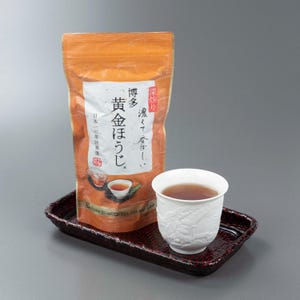 May include: A stand-up pouch of Golden Roasted Tea with orange packaging and Japanese text, placed on a dark red rectangular woven tray next to a white ceramic teacup filled with dark brown tea. The pouch features illustrations of tea.