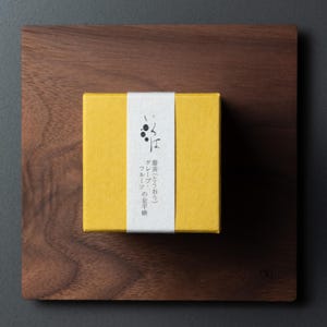 May include: A yellow box with Japanese text and a white band, placed on a dark wooden square. The box likely contains food. The wooden square has a natural wood grain. The box is likely a food product.
