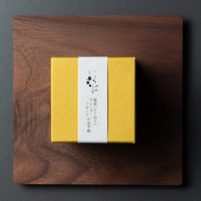 May include: A yellow box with Japanese text and a white band, placed on a dark wooden square. The box likely contains food. The wooden square has a natural wood grain. The box is likely a food product.
