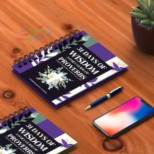 May include: Two purple and green spiral bound journals with the text "31 Days of Wisdom A Daily Guide of Proverbs" on the cover. The journals are on a wooden surface with a pen and a smartphone.