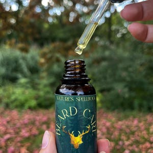 Op de afbeelding: Een bruine glazen fles baardolie met een pipet. Het etiket luidt "NATURE'S SPELLBOOK BEARD CARE" met een gouden hertenillustratie. De pipet is gevuld met een gele vloeistof en wordt boven de fles gehouden.