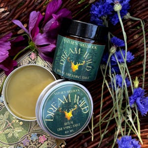 May include: An overhead shot of two containers of ritual dreaming salve, one in a jar and one in a tin, with the text "Dream Wild" and "Nature's Spellbook". The image is surrounded by purple and blue flowers.