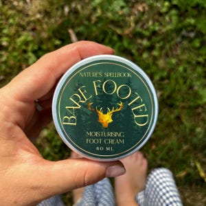 Könnte beinhalten: Eine runde, grün-goldene Dose "Bare Footed" Feuchtigkeitscreme für die Füße. Das Etikett zeigt eine goldene Hirschsilhouette und den Text "Nature's Spellbook". Der Behälter wird in einer Hand vor einem verschwommenen grünen Hintergrund gehalten.