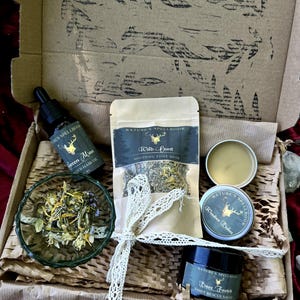 Op de afbeelding: Een open kartonnen doos gevuld met zelfzorgproducten. De doos bevat een glazen kom met gedroogde kruiden, een fles Green Man olie, een zakje Wild Hunt voetbad en blikken Wonder Balm en Bare Footed voetreddingszalf.