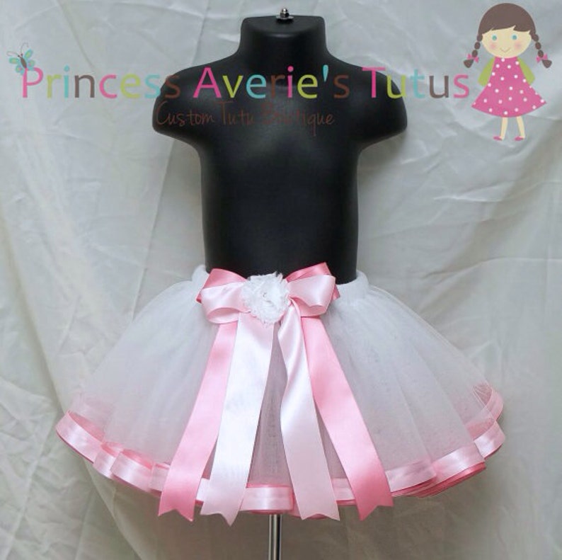May include: White tulle tutu skirt with a pink satin ribbon bow and a white flower accent. The skirt has a pink satin trim around the bottom.