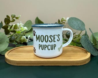 May include: White enamel mug with a green rim and a green inscription that reads "Moose's Pupcup". The mug is decorated with brown paw prints and a green bone.