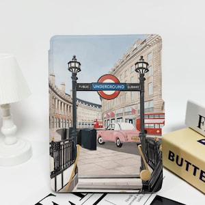 May include: A decorative case featuring a London Underground scene with a pink car, red double-decker bus, and the text "UNDERGROUND SUBWAY." The case has a black metal fence and a vintage street lamp.
