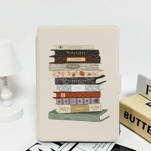Puede incluir: Una funda beige para lector de libros electrónicos con una pila de libros ilustrados. Los títulos de los libros incluyen "Midnights", "Evermore", "Folklore", "Reputation", "1989" y "Fearless". La funda tiene un recorte para el lector y un fondo blanco.