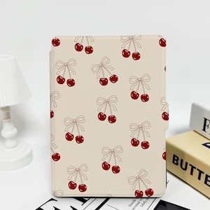 Puede incluir: Una funda beige para lector electrónico Kindle Paperwhite con un patrón de cerezas rojas y lazos. La funda tiene un fondo blanco con un patrón repetitivo de cerezas rojas y lazos blancos.