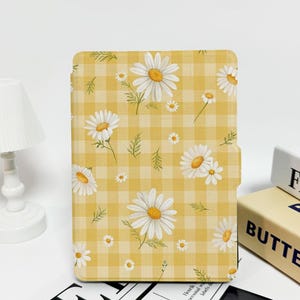 Puede incluir: Una funda para tableta con un diseño de margaritas amarillas y blancas. La funda presenta un fondo de cuadros vichy con margaritas blancas y hojas verdes. La funda está cerrada y parece estar hecha de un material duradero. El tema general es floral y primaveral.