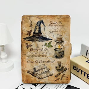 May include: A decorative journal with a vintage, parchment-like appearance. The cover features illustrations of a witch's hat, potion bottles, and an open book, with handwritten text and botanical details. A small white lamp is visible on the left.