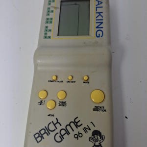 May include: A beige handheld electronic game console with a vertical LCD screen, yellow control buttons, and the text TALKING, BRICK GAME 96 IN 1, and E-23 PLUS MARK II printed on the front. The device features a small graphic of a person.