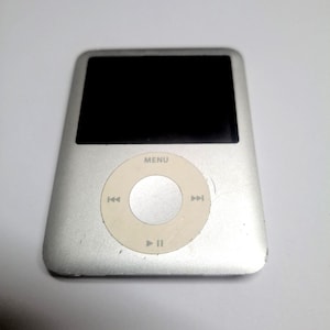 May include: A silver digital music player with a rectangular black screen and a circular white click wheel. The click wheel features a central silver button and icons for menu, play, pause, and track skipping. The device has a metallic finish.