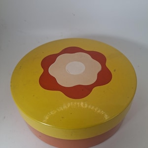 May include: A round metal storage tin with a yellow lid featuring a large, stylised flower graphic in orange and cream. The base of the container is a solid, muted orange colour. The lid is flat and circular, while the body of the tin is cylindrical.