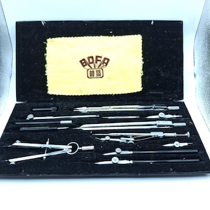 May include: A black rectangular case containing a set of silver metal drafting compasses and technical drawing tools. The interior features a yellow cloth patch with the text BOFA and Chinese characters. The tools include various dividers.