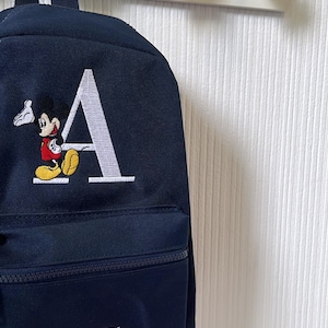 May include: Navy blue backpack featuring a white embroidered letter "A" and a Mickey Mouse design. The front pocket displays the name "ASHTON" in white. The backpack hangs on a white rack with star-shaped hooks, suitable for a child's room.