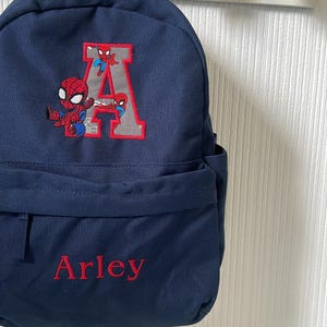 May include: A navy blue backpack with a Spider-Man design, the letter "A", and the name "Arley" embroidered in red. It has a front pocket and black straps. The bag hangs on a white wall with star-shaped hooks.