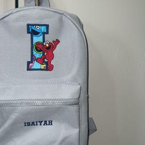 May include: A pale grey backpack with a front pocket and a top handle. The backpack has the name "ISAIYAH" in navy blue lettering and a colourful embroidered design of the letter "I" with Sesame Street characters.