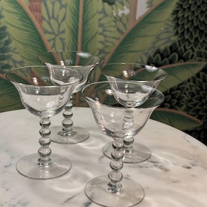 May include: Set of four clear glass stemmed cocktail glasses with beaded stems. The glasses are arranged on a white marble surface.