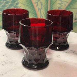 May include: Three ruby red vintage drinking glasses. Each glass has a faceted, geometric design on the lower portion and a smooth, cylindrical upper section. The glasses are made of a dark, transparent glass.