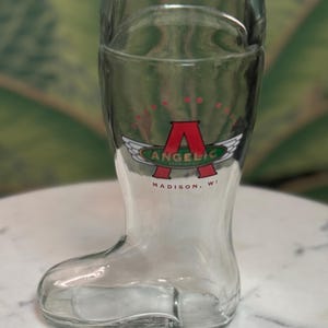 May include: Clear glass boot-shaped beer glass with the word "ANGELIC" in green and white with a red "A" and wings logo. The text "MADISON, WI" is below the logo. The glass is empty and sits on a white marble surface.