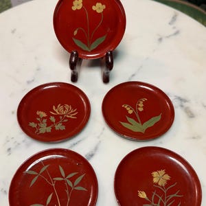 May include: Set of five red lacquered coasters with floral designs. One coaster is displayed on a wooden stand. The coasters feature various flowers in gold and green, including chrysanthemums and lilies.