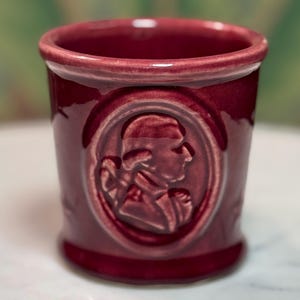 May include: A burgundy ceramic tumbler with a raised relief portrait of a person in an oval frame. The tumbler has a cylindrical shape with a slightly flared rim and a base. The background is blurred.