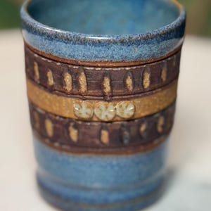 May include: A handmade ceramic tumbler with a blue interior and rim. The exterior features bands of brown and tan with decorative accents. The tumbler is approximately 4 inches tall and 3 inches in diameter.