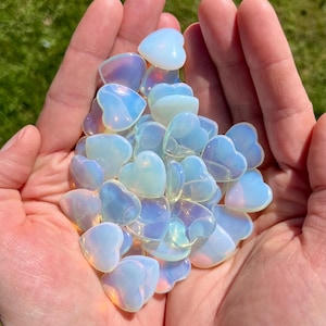 Può includere: Una manciata di pietre a forma di cuore opalescenti nelle tonalità del bianco, del blu e del viola. Le pietre traslucide sono tenute in mani aperte su uno sfondo di erba verde. I cuori variano per dimensioni e forma.