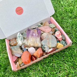 Pode incluir: Uma caixa rosa cheia de uma variedade de cristais e pedras coloridas, incluindo ametista, quartzo rosa e ágata. A caixa está sentada em um leito de grama verde. "Obrigado por seu pedido" está impresso em um adesivo na caixa.