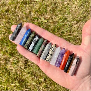May include: A collection of fifteen small, polished crystal points in various colours and patterns, including black, pink, blue, green, white, purple, orange, and brown, arranged in a row across an open palm against a blurred green background.