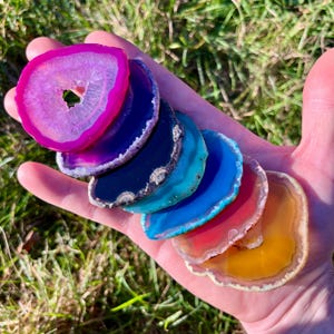 Könnte beinhalten: Eine Hand hält eine Sammlung farbenfroher Achatscheiben. Die Scheiben sind fächerförmig angeordnet und zeigen eine Vielzahl von Farben, darunter Rosa, Lila, Blau, Türkis, Rot und Gelb. Jede Scheibe hat ein einzigartiges Muster und eine polierte Oberfläche.