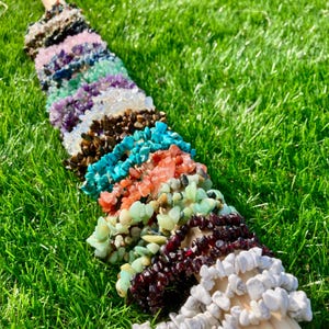 Peut inclure: Un ensemble de bracelets en pierres précieuses colorées, chacun composé de différents types de pierres, notamment turquoise, cornaline et agate blanche. Les bracelets sont disposés en rangée sur un lit d'herbe verte.