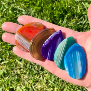 Può includere: Una mano che tiene una collezione di pietre di agata lucidata in vari colori. Le pietre includono sfumature di rosso, marrone, viola, verde e blu, ognuna con motivi a bande unici. Le pietre hanno la forma di una goccia e sono esposte su uno sfondo di erba verde.