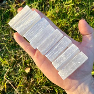 Op de afbeelding: Een hand houdt verschillende rechthoekige, witte selenietkristallen staven vast. De staven zijn in een rij gerangschikt en tonen hun natuurlijke, getextureerde oppervlak. De achtergrond is wazig groen gras.