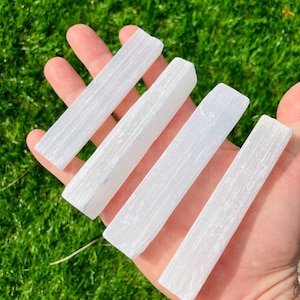 Op de afbeelding: Vier witte selenietkristallen staven, elk rechthoekig met een textuuroppervlak. De staven worden in een open hand gehouden tegen een achtergrond van groen gras.