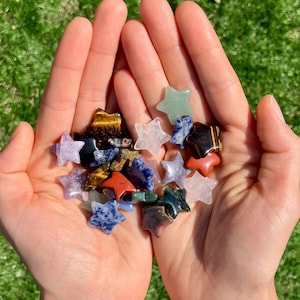 May include: A selection of star-shaped gemstones in diverse colours, such as purple, blue, green, and clear. The gemstones are held in open hands against a background of green grass. The gemstones appear polished and smooth.