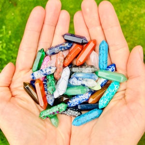 May include: A collection of colorful crystal wands in various shades of blue, green, orange, pink, and white. The crystals are held in open hands against a blurred green background. Each wand is approximately 2-3 inches long.