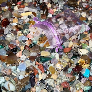 May include: A close-up of a large collection of colourful, polished stones and pebbles. The stones are of various shapes and sizes, and some are arranged in a small purple mesh bag.