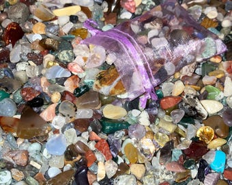 May include: A close-up of a large collection of colorful, polished stones and pebbles. The stones are of various shapes and sizes, and some are arranged in a small purple mesh bag.