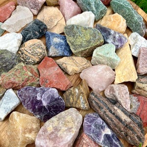 May include: A selection of raw gemstones in a variety of colours, including red, purple, green, and beige. The stones vary in size and shape, with rough, natural textures. The gemstones are arranged on a wooden surface.