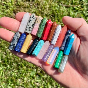 May include: A selection of crystal wands in a variety of colours and patterns, including white, black, blue, yellow, green, red, and purple. The crystals are held in a hand against a backdrop of green grass.