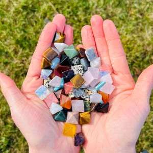 May include: A collection of small, pyramid-shaped gemstones in various colours, including yellow, brown, green, pink, and blue. The gemstones are held in open hands, with a blurred green grass background.