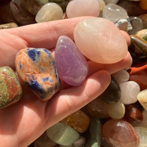 May include: A collection of tumbled stones in various colours, including purple amethyst, pink rose quartz, and blue and orange sodalite. The stones are smooth and polished, and they are arranged in a hand.