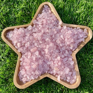 May include: A wooden star-shaped bowl filled with small, light pink rose quartz crystals. The crystals are irregularly shaped and vary in size. The bowl sits on green grass, creating a natural and earthy aesthetic.