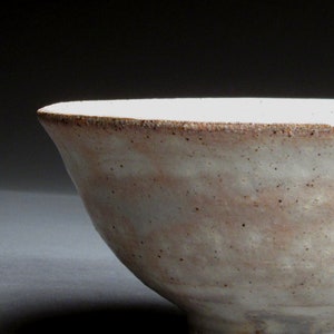 May include: A handcrafted ceramic bowl with a speckled, off-white glaze. The rim has a darker, textured edge. The bowl's shape is slightly flared, and the exterior shows subtle variations in color, suggesting a natural, organic aesthetic.